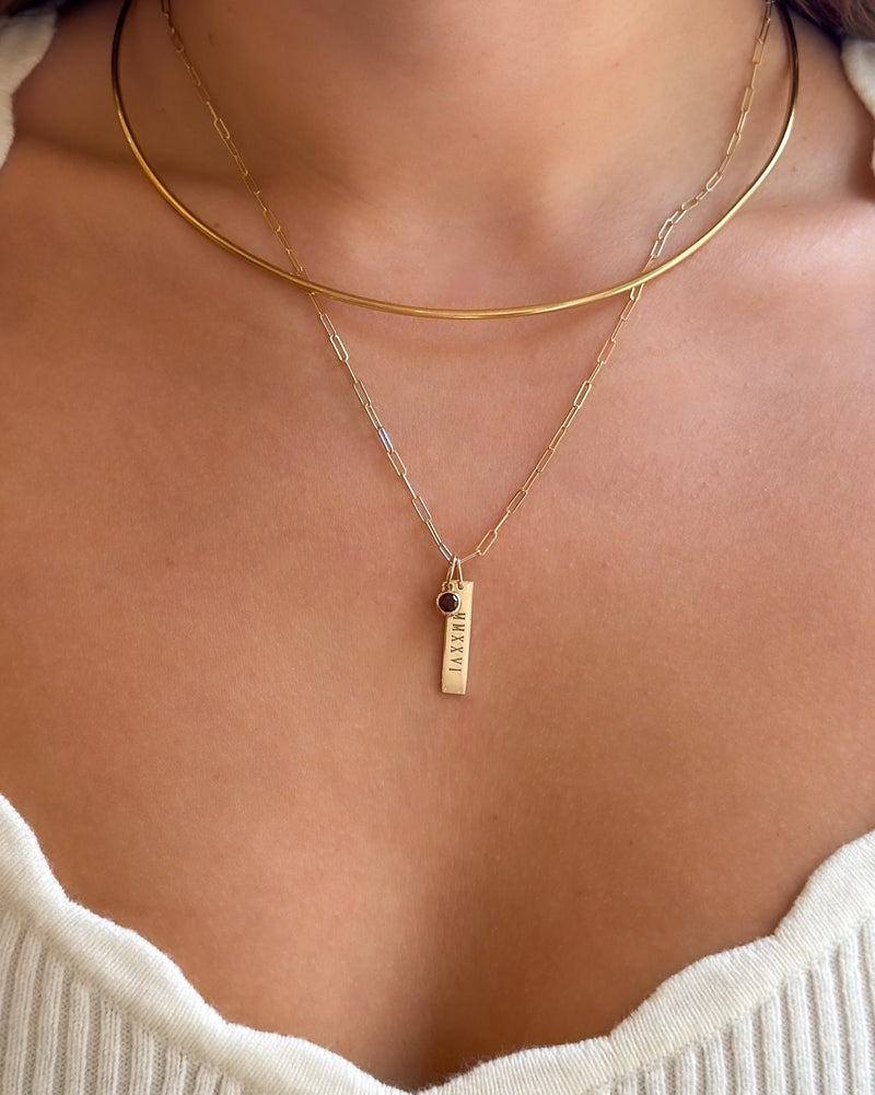 A close-up of a person's neck and chest wearing a layered gold necklace set. The set includes a thin circular choker and a delicate chain necklace with a small vertical bar pendant featuring a tiny dark stone accent.