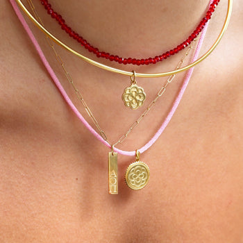 A close-up of a person's neck and collarbone wearing a layered necklace set. The set includes a red beaded choker, a gold bangle, a delicate gold chain with a small flower charm, a pink cord necklace with a gold bar charm, and a pink cord necklace with a gold medallion charm.