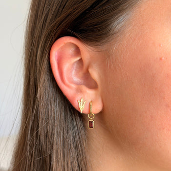 A close-up of a person's ear wearing a pair of small gold earrings, one with a gold leaf design and the other with a gold hoop and a small dark red rectangular drop, against a soft light background.