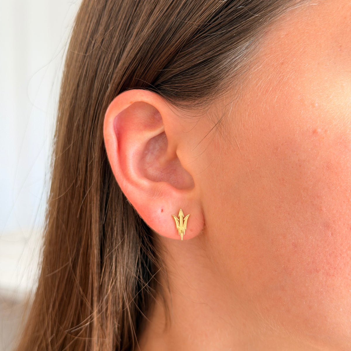 A close-up side view of a woman's ear wearing a small gold trident stud earring.