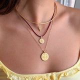A close-up of a model's neck and chest wearing a layered necklace set. The set includes a gold chain choker with a small lightning bolt pendant, a dark red cord necklace with a round gold coin pendant, a gold chain necklace with a larger round gold coin pendant, and a delicate gold chain.
