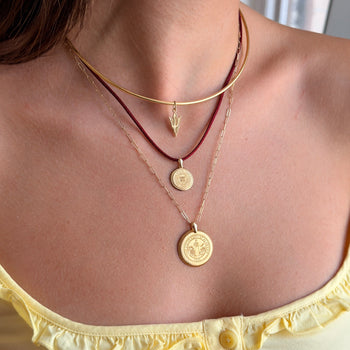 A close-up of a model's neck and chest wearing a layered necklace set. The set includes a gold chain choker with a small lightning bolt pendant, a dark red cord necklace with a round gold coin pendant, a gold chain necklace with a larger round gold coin pendant, and a delicate gold chain.