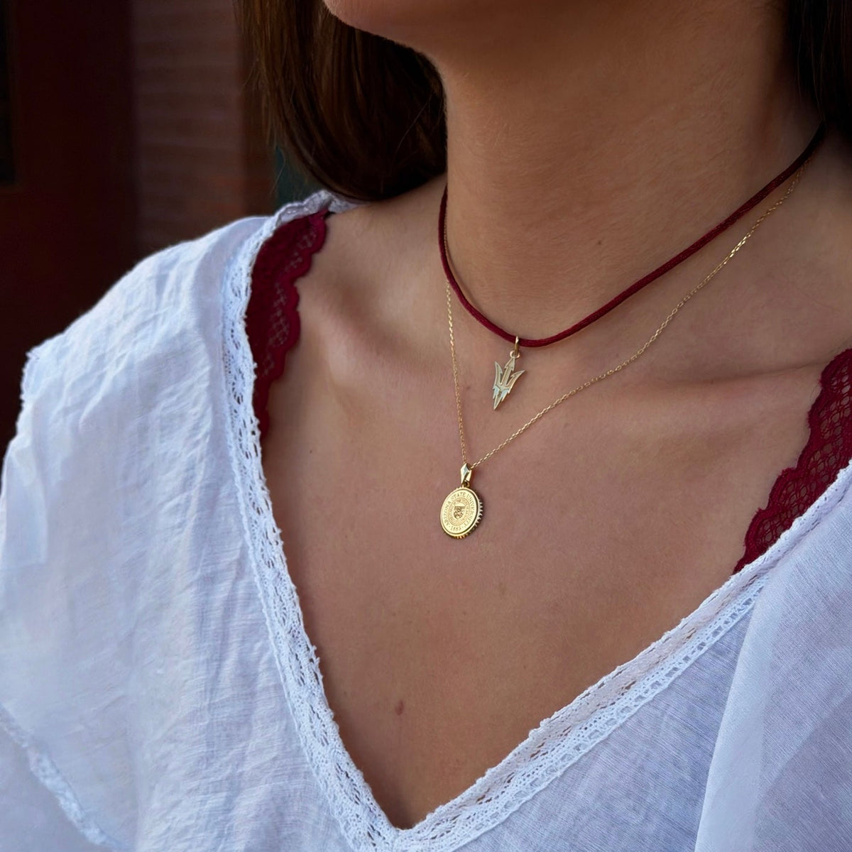 A close-up of a person wearing layered necklaces, including a gold chain with a round pendant and a gold chain with a leaf pendant over a dark red cord necklace, paired with a white lace-trim blouse.