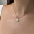 A close-up of a model's neck and chest wearing a delicate gold necklace with a round pendant. The necklace has a 16-inch ball chain and a small circular pendant engraved with the letter 'S'.