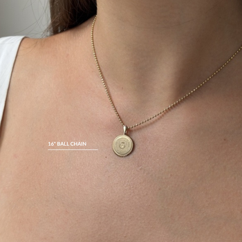 A close-up of a model's neck and chest wearing a delicate gold necklace with a round pendant. The necklace has a 16-inch ball chain and a small circular pendant engraved with the letter 'S'.