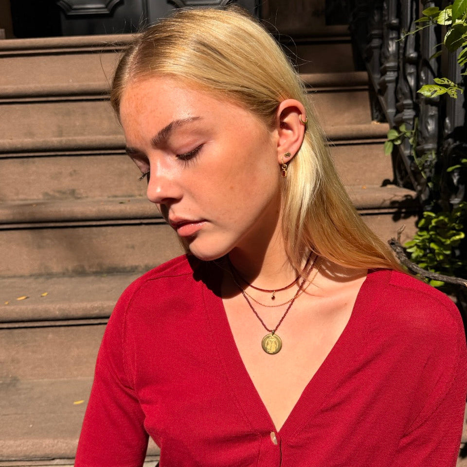 A blonde woman wearing a red button-up cardigan and layered necklaces, sitting on stone steps in outdoor sunlight.
