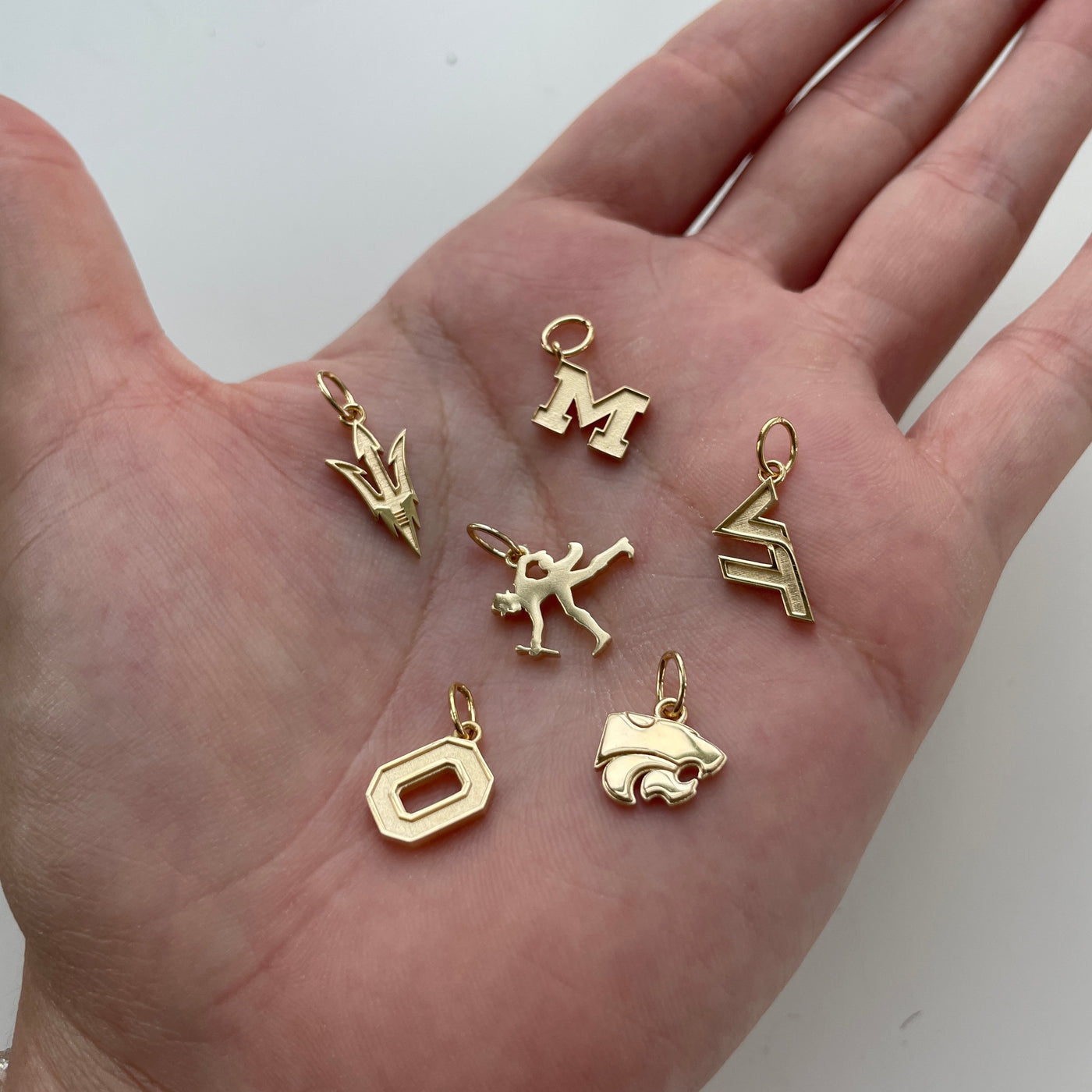 Michigan Logo Charm, Virginia Tech Logo Charm, Kansas State Wildcat Charm, Ohio State Logo Charm, ASU Pitchfork Charm, Wake Forest Walking Deacon Charm shown in gold in hand