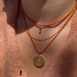 Close-up of a person wearing layered necklaces, including an orange cord necklace with a round gold pendant, a thin gold chain, and an orange beaded necklace, over a peach knit top.