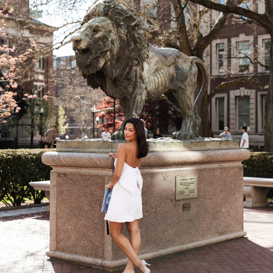 Woman in a white dress standing next to a lion statue in an urban park setting.
