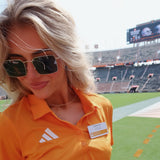 A woman wearing an orange Adidas polo shirt and sunglasses stands on a football field with stadium seating in the background.