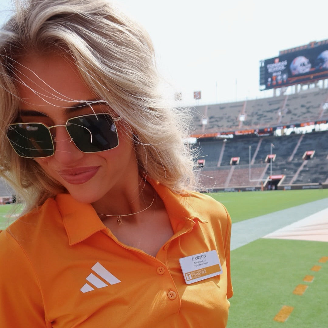 A woman wearing an orange Adidas polo shirt and sunglasses stands on a football field with stadium seating in the background.