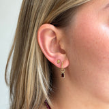 A close-up of a woman's ear wearing a gold huggie hoop earring with a small gold cartilage stud and a dangling gold hook with a rectangular dark red gemstone.