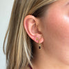 A close-up of a woman's ear wearing a gold huggie hoop earring with a small gold cartilage stud and a dangling gold hook with a rectangular dark red gemstone.