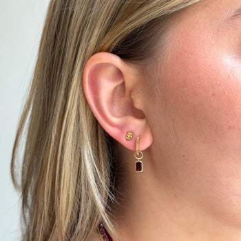 A close-up of a woman's ear wearing a gold huggie hoop earring with a small gold cartilage stud and a dangling gold hook with a rectangular dark red gemstone.