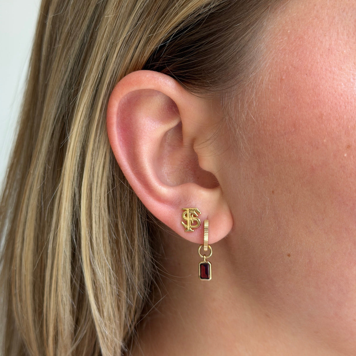 A close-up of a woman's ear wearing a gold huggie hoop earring with a small drop charm featuring a rectangular dark red gemstone.