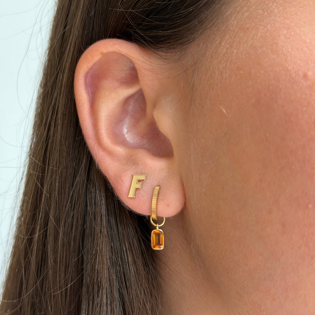 Close-up of a person's ear wearing a small gold initial 'F' stud earring and a gold huggie hoop with a dangling rectangular amber gemstone.