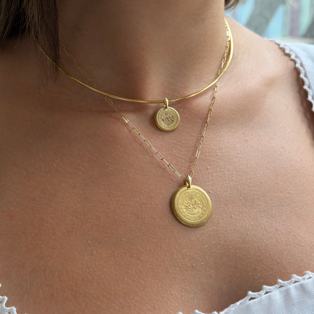 A close-up of a model wearing a layered gold necklace set, featuring a thin curved chain with a small round pendant and a longer chain with a larger round medallion pendant.