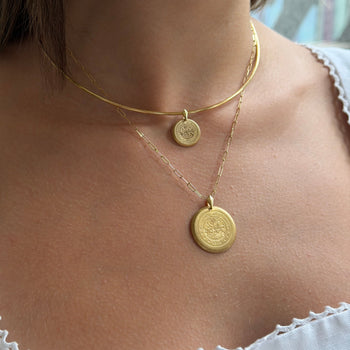 A close-up of a model wearing a layered gold necklace set, featuring a thin curved chain with a small round pendant and a longer chain with a larger round medallion pendant.