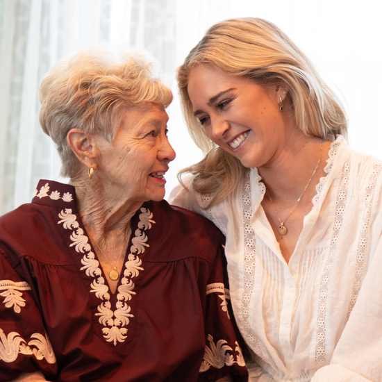 Two women, one elderly and one younger, smiling and looking at each other against a white curtain background.