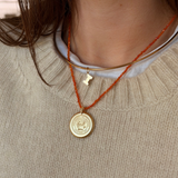 A close-up of a person wearing a layered necklace set. The set includes a gold bangle collar, a gold initial 'I' pendant, and a gold round medallion pendant on an orange cord, layered over a white shirt and a beige knit sweater.