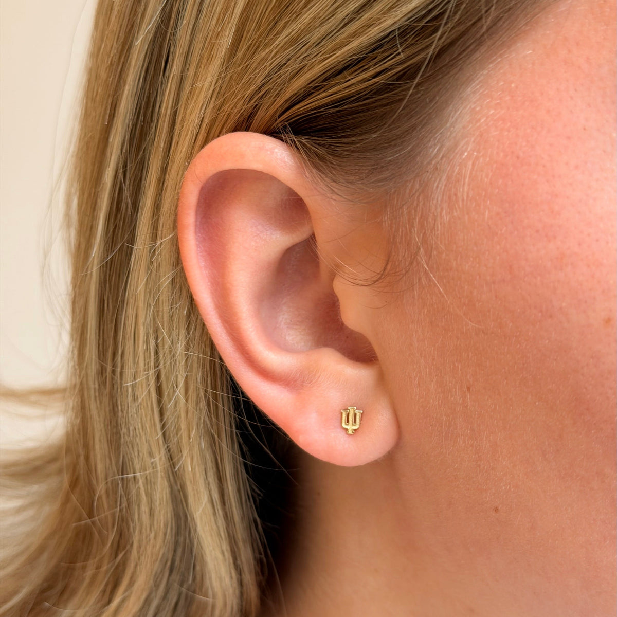 A close-up of a person's ear wearing a tiny gold stud earring with a small stylized symbol, against a soft beige background.