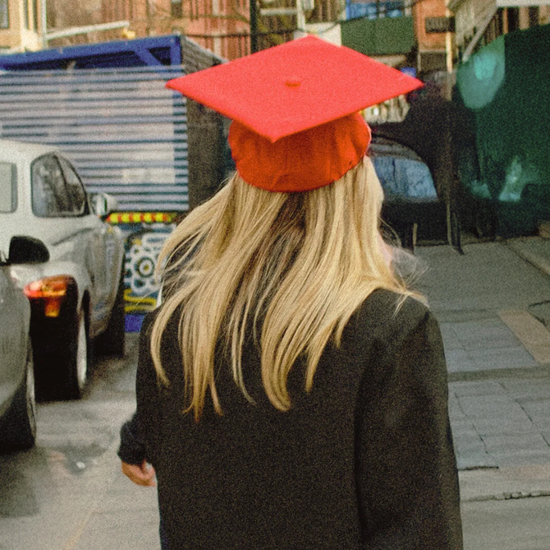 Person wearing a red graduation cap on a street with cars and buildings in the background
