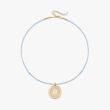 A delicate light blue cord necklace with a gold-toned circular pendant and extension chain, displayed on a clean off-white background.