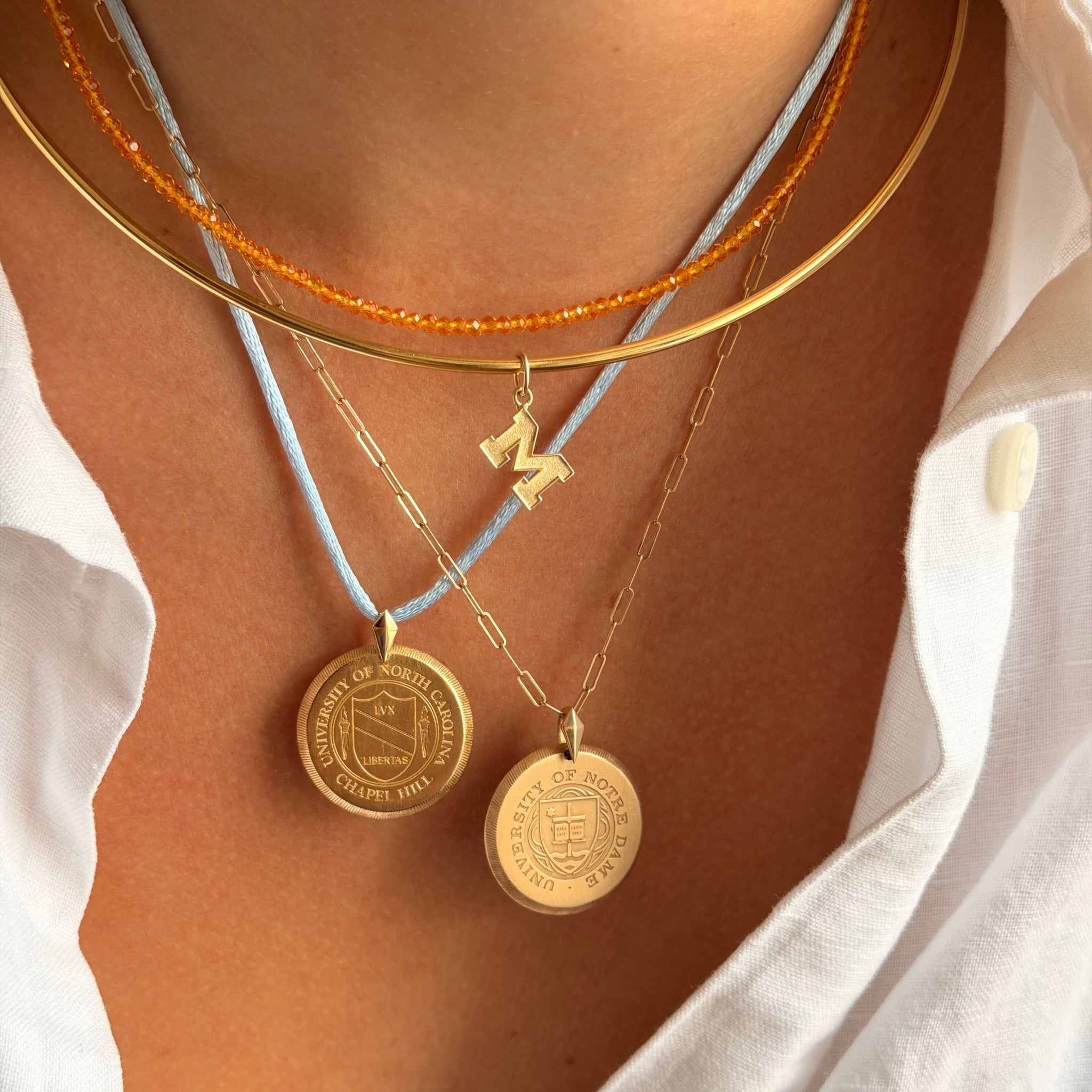 Close-up of a person's neck and chest wearing layered necklaces: a gold chain with a small gold initial pendant, a gold bangle, a gold chain with round medallion pendants on light blue cords, and an orange beaded necklace.