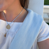 A close-up lifestyle shot of a woman wearing a layered necklace set featuring a gold coin pendant, a thin gold chain, and a light blue cord necklace, along with a pearl stud earring.