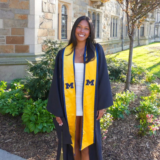 Graduate in a yellow and blue gown standing outdoors with a building in the background
