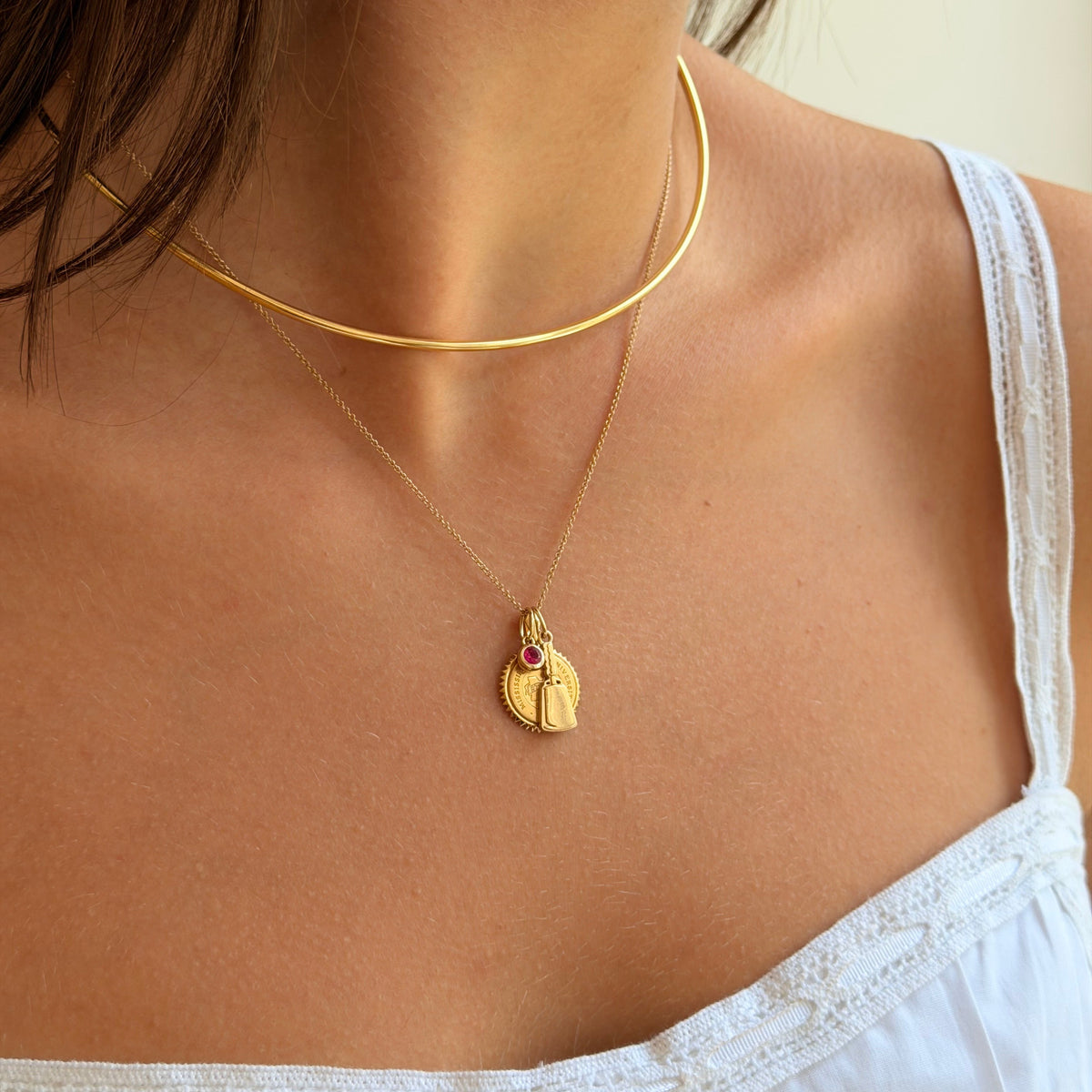 A close-up of a model wearing a double-layered gold necklace. The top layer is a thin curved cable choker, and the bottom layer is a delicate chain with a small gold pendant featuring a red gemstone accent.