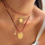 A close-up of a person's neck and chest wearing a layered necklace set. The set includes a delicate pinkish-red beaded chain, a thin gold chain, a red cord with a gold coin pendant, and a gold chain with a small gold tag charm.