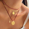 A close-up of a person's neck and chest wearing a layered necklace set. The set includes a delicate pinkish-red beaded chain, a thin gold chain, a red cord with a gold coin pendant, and a gold chain with a small gold tag charm.