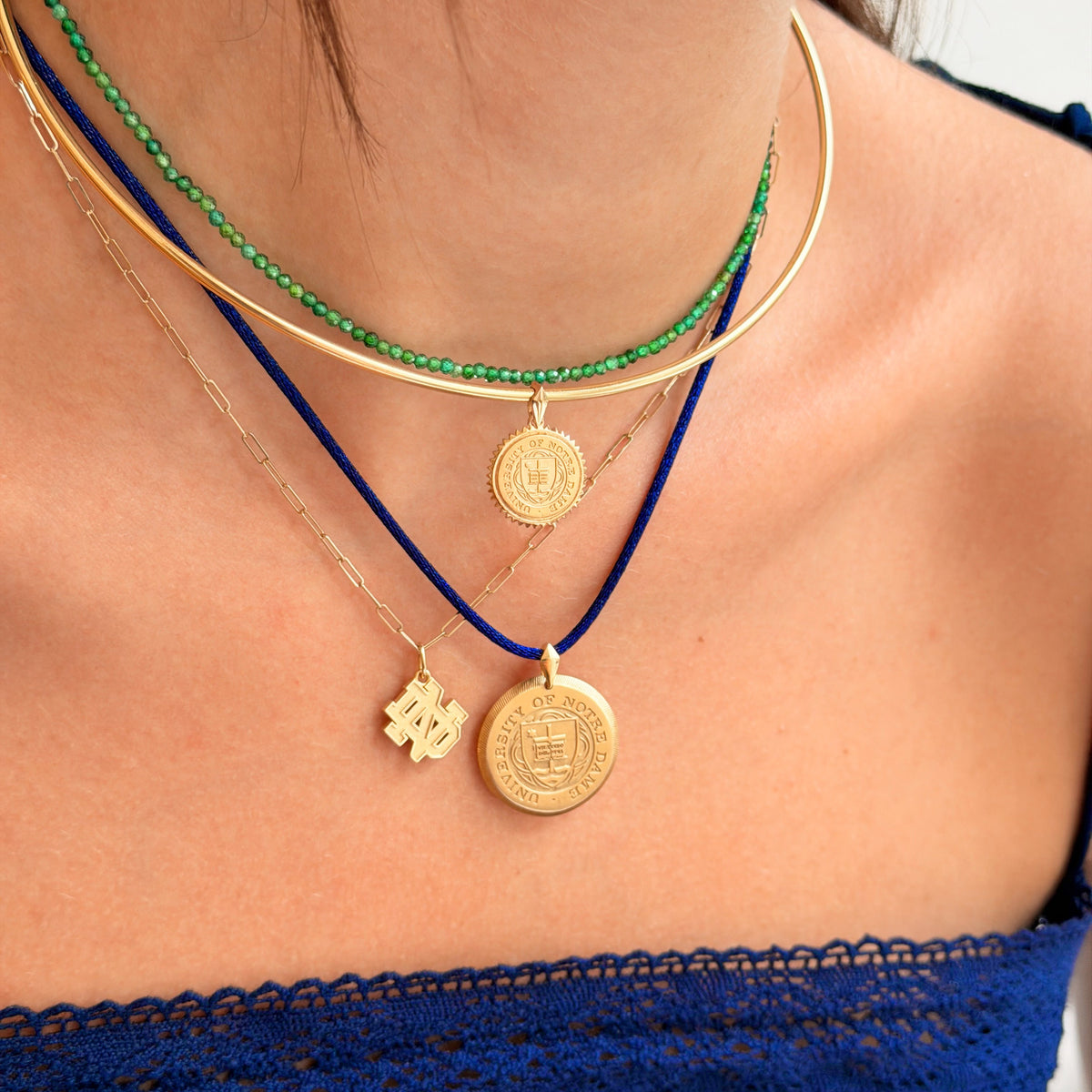 A close-up of a person's neck and chest wearing layered necklaces: a green beaded choker, a gold bangle, a gold chain with a round coin-style pendant, a gold chain with a small dangling charm, and a dark blue cord necklace with a round gold coin pendant.