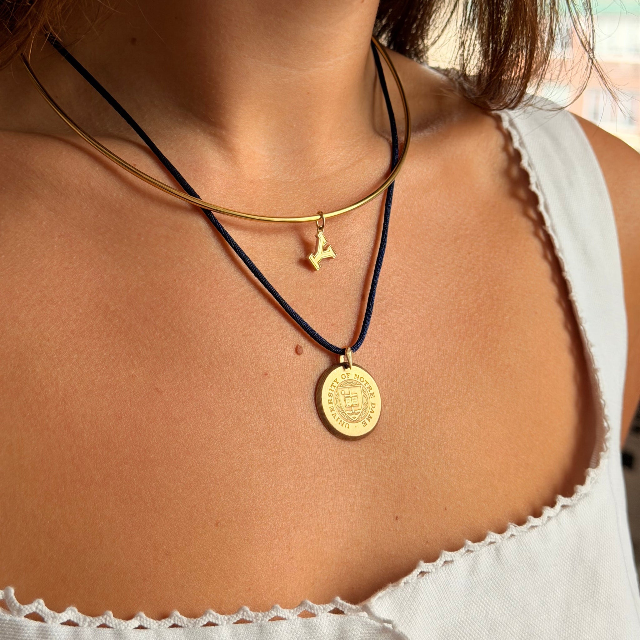 A close-up of a person wearing layered necklaces: a gold chain with a small gold anchor charm, a gold coin pendant necklace, and a thin black cord necklace. The person is wearing a white top.
