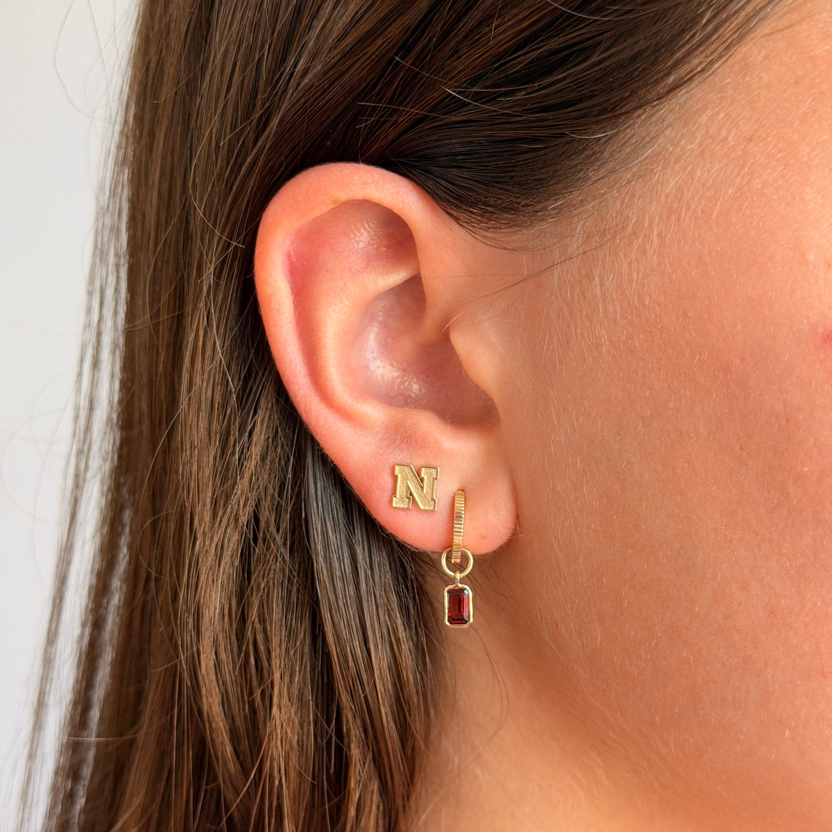 A close-up of a person's ear wearing a gold hoop earring with a small gold initial 'N' stud and a dangling gold barrette with a rectangular red gemstone.