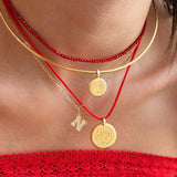 A close-up of a person wearing layered necklaces: a red beaded choker, a gold bangle, a gold chain with a coin pendant, a red cord necklace with a gold coin pendant, and a small gold initial 'N' charm. The person is wearing a red off-the-shoulder top.