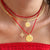 A close-up of a person wearing layered necklaces: a red beaded choker, a gold bangle, a gold chain with a coin pendant, a red cord necklace with a gold coin pendant, and a small gold initial 'N' charm. The person is wearing a red off-the-shoulder top.