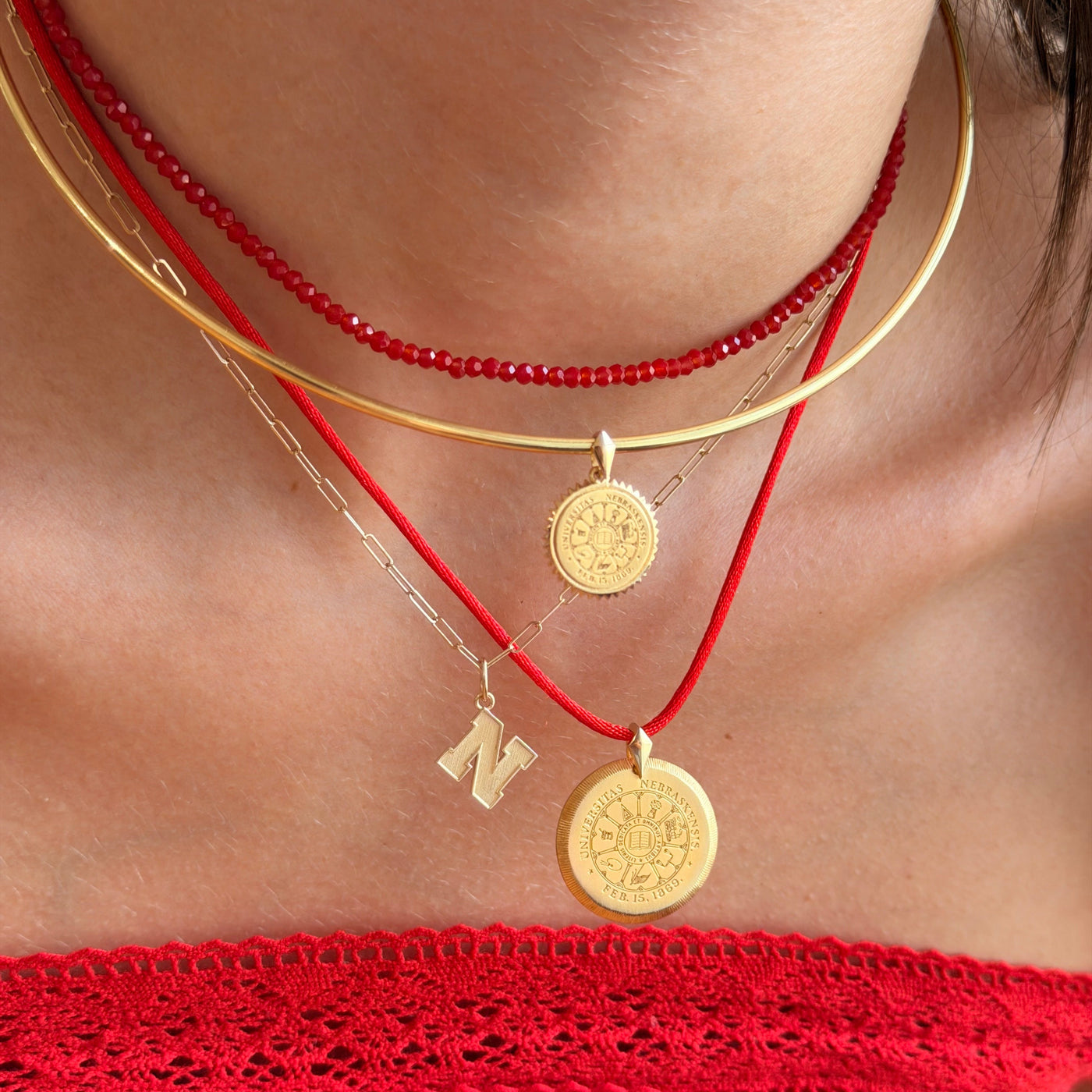 A close-up of a person wearing layered necklaces: a red beaded choker, a gold bangle, a gold chain with a coin pendant, a red cord necklace with a gold coin pendant, and a small gold initial 'N' charm. The person is wearing a red off-the-shoulder top.