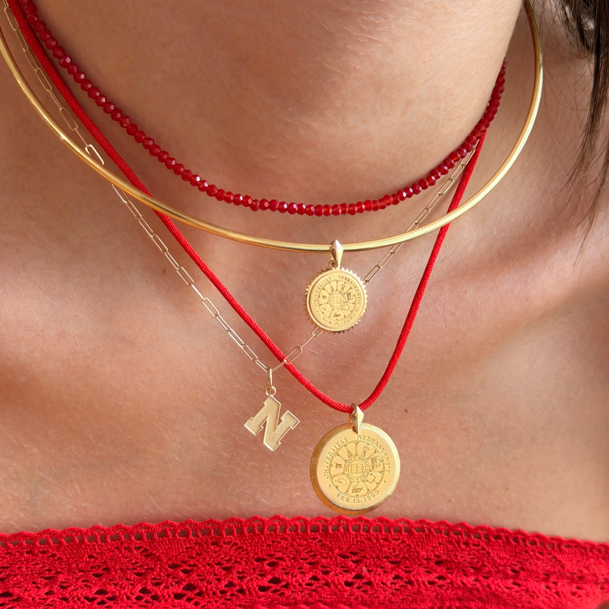 A close-up of a person wearing layered necklaces: a red beaded choker, a gold bangle, a gold chain with a coin pendant, a red cord necklace with a gold coin pendant, and a small gold initial 'N' charm. The person is wearing a red off-the-shoulder top.
