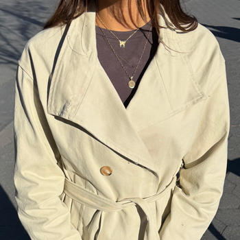 A person outdoors wearing a light beige belted double-breasted jacket over a dark purple top and blue jeans, holding a brown suede clutch bag with a flower charm.