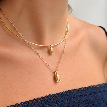A close-up of a model's neck and upper chest wearing two layered gold necklaces: a thin chain necklace with a small Buddha head pendant and a slightly thicker curved chain necklace with a larger Buddha head pendant.