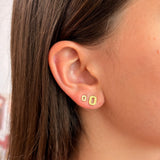 A close-up of a person's ear wearing two small gold geometric stud earrings, with brown hair and part of the face visible.