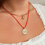 A close-up of a person's neck and chest wearing two layered necklaces: a thin gold bangle with a small dangling charm and a red cord necklace with a round gold medallion pendant.