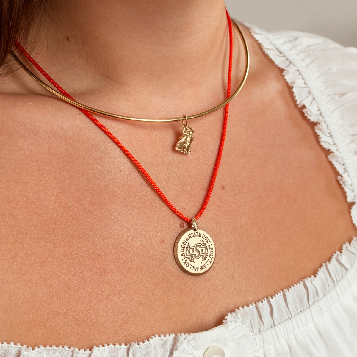 A close-up of a person's neck and chest wearing two layered necklaces: a thin gold bangle with a small dangling charm and a red cord necklace with a round gold medallion pendant.