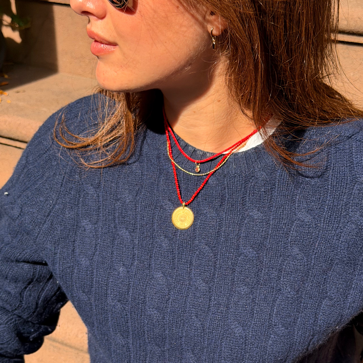 A woman wearing a navy blue cable-knit crewneck sweater, styled with light-wash blue jeans, a black belt, and layered red cord necklaces, sitting outdoors on stone steps.