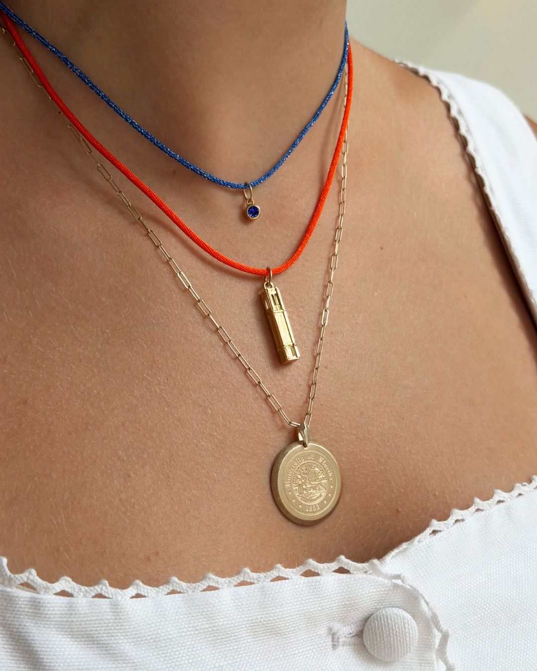 A close-up of a person's neck and chest wearing three layered necklaces: a blue beaded cord necklace with a small blue stone pendant, an orange cord necklace with a gold cylindrical pendant, and a gold chain necklace with a round gold coin pendant.