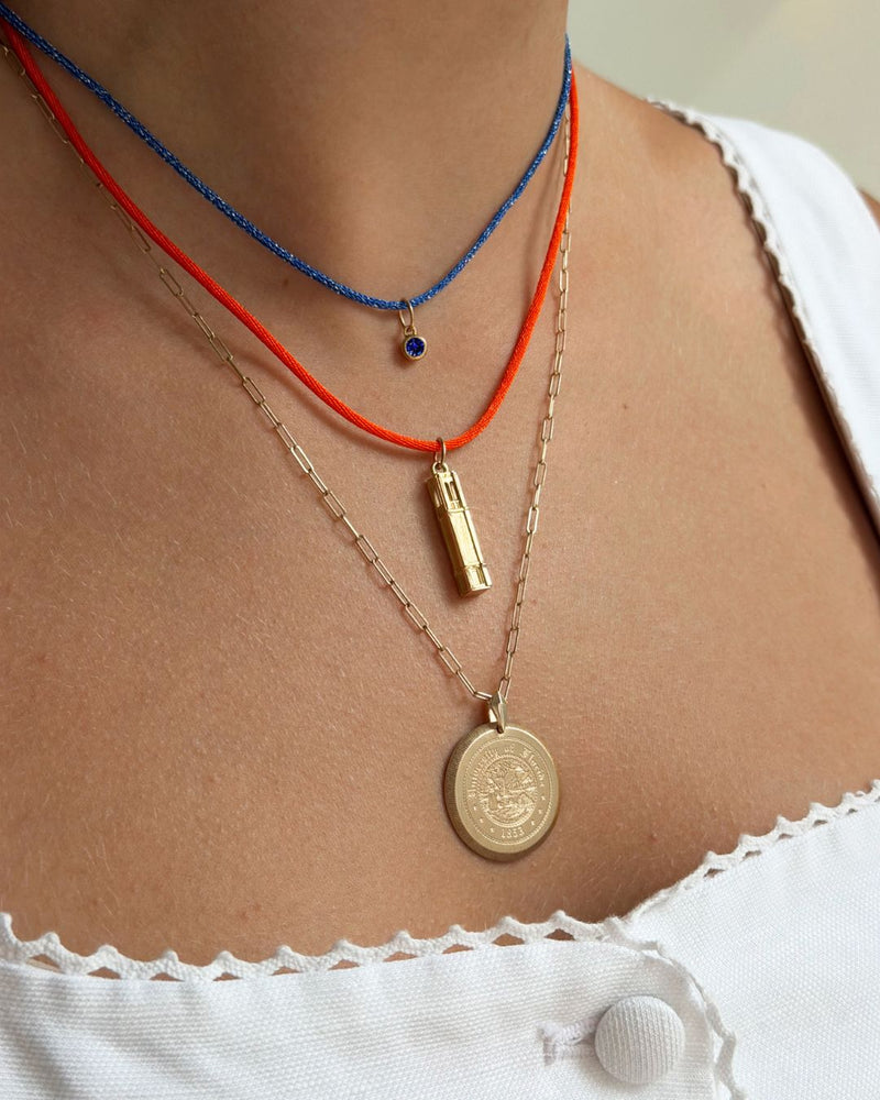 A close-up of a person's neck and chest wearing three layered necklaces: a blue beaded cord necklace with a small blue stone pendant, an orange cord necklace with a gold cylindrical pendant, and a gold chain necklace with a round gold coin pendant.