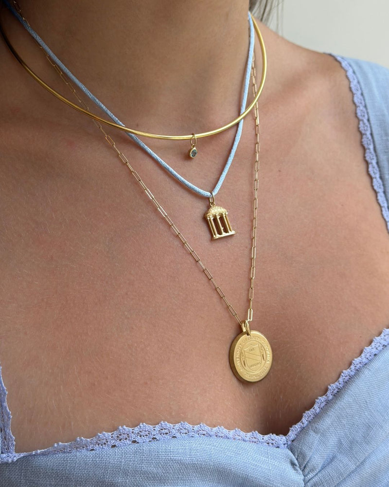 A close-up of a person's neck and chest wearing layered necklaces: a gold chain with a small birdcage pendant, a gold chain with a round coin pendant, and a light blue cord necklace with a tiny green stone accent.
