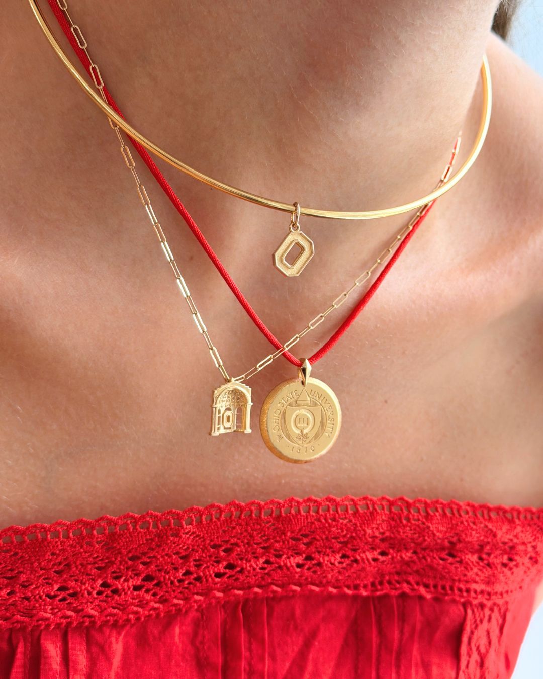 A close-up of a person wearing layered necklaces: a gold chain choker with a small geometric pendant, a gold chain with a small bird pendant, and a red cord necklace with a round gold medallion pendant.